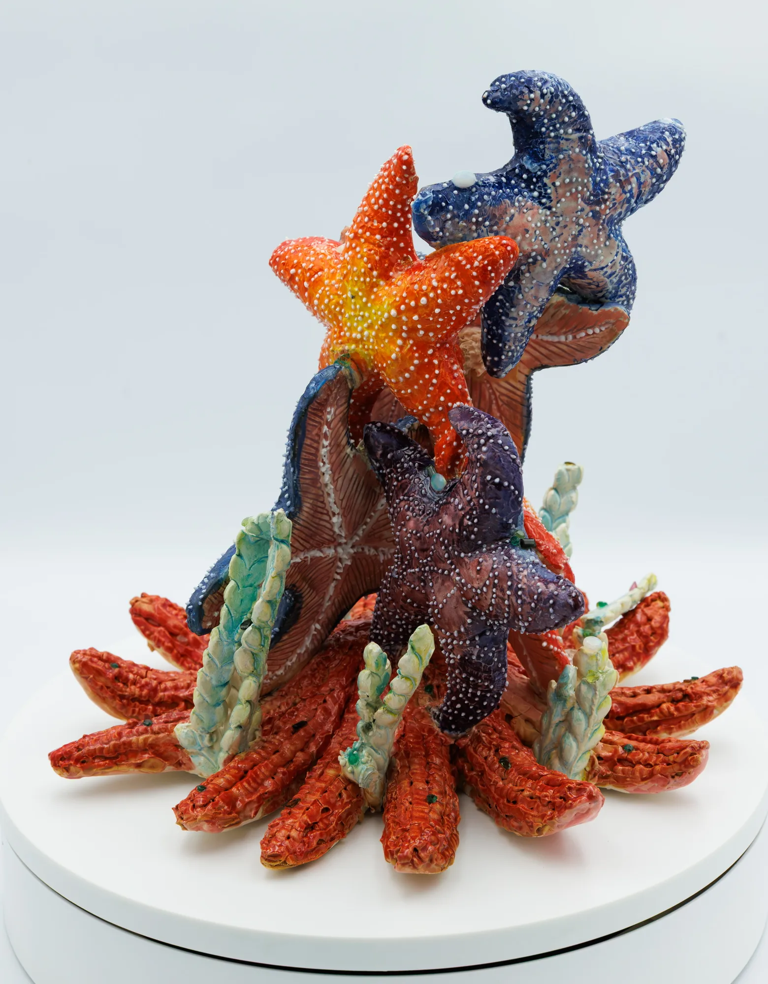 Glazed porcelain sculpture of dotted orange, blue and purple starfish rising above a ring of red ridged arms and pale green fronds, on a white stand.